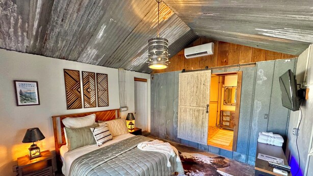 Quarto com cama, mesa de cabeceira, luminária, televisão presa na parede e ventilador de teto.