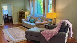 Sala de estar com sofá, um banquinho e uma mesinha com abajur. Estante e um espaço de refeições são visíveis ao fundo.