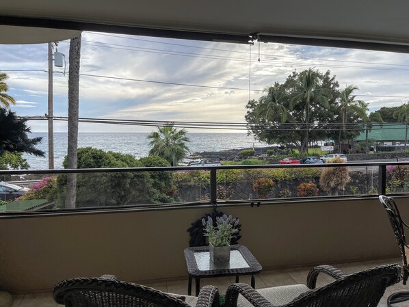 A balcony with a view of the ocean, a small table with a potted plant, wicker furniture, and a stone wall. - Fabulous Ocean Views : Whales : Dolphins : Big Island Fun! (Kailua-Kona)