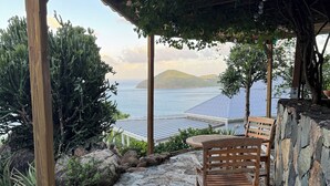 A stone patio with a wooden pergola and a wooden bench overlooking a body of water and distant hills.