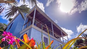 A blue building with a balcony, surrounded by tropical plants and flowers.