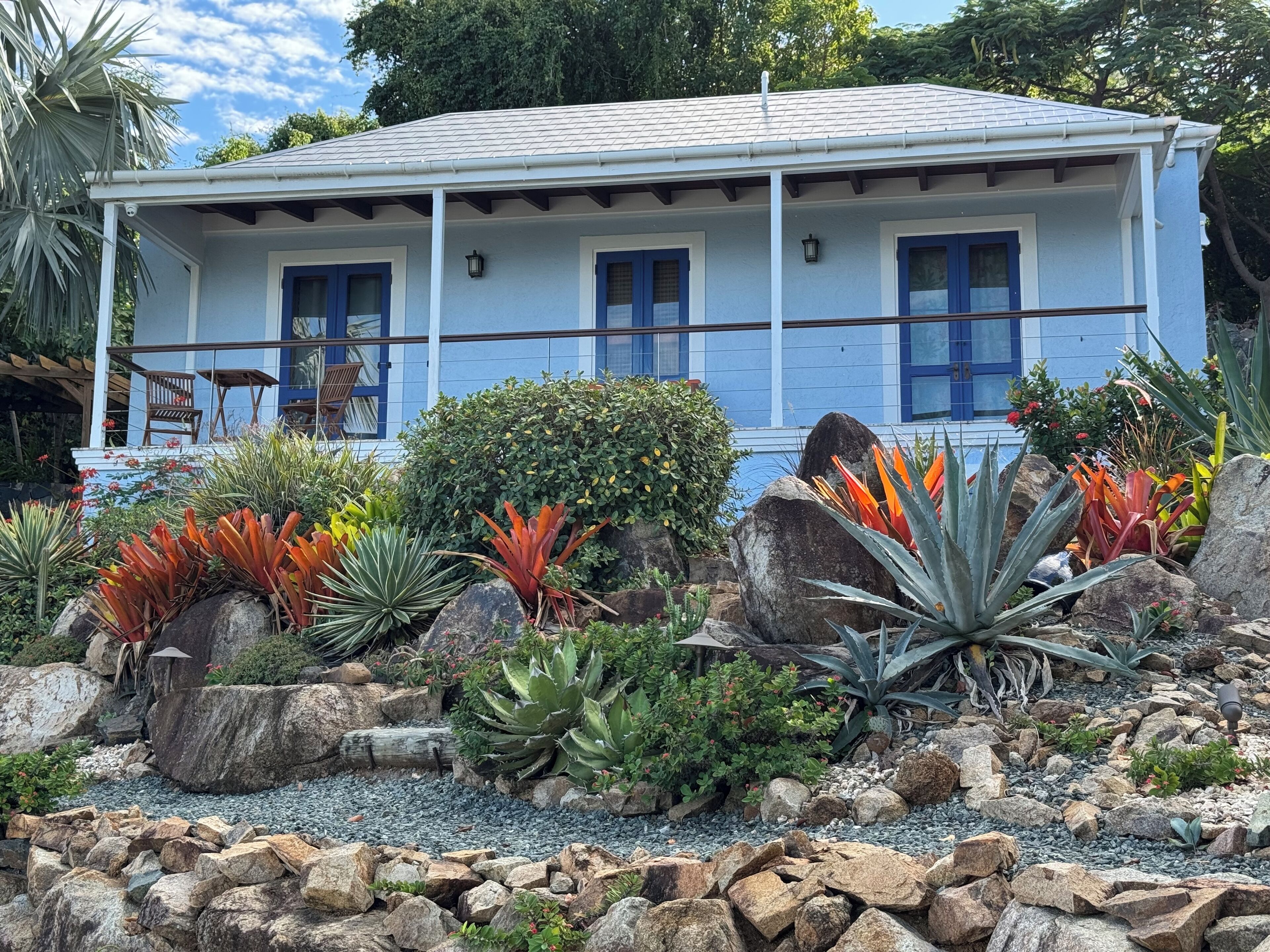 A house with a blue exterior, a covered porch with chairs, and a garden with various plants and rocks.