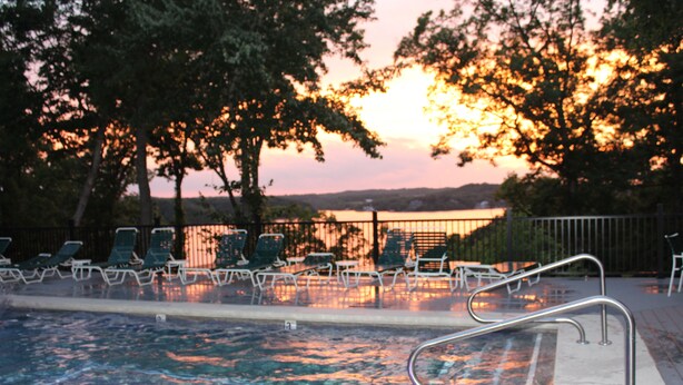 A swimming pool with metal railings and lounge chairs, surrounded by trees and a sunset in the background.
