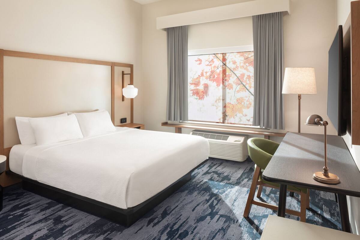 Une chambre d’hôtel avec un grand lit, un bureau avec une lampe, une chaise, une télévision et une fenêtre avec des rideaux.