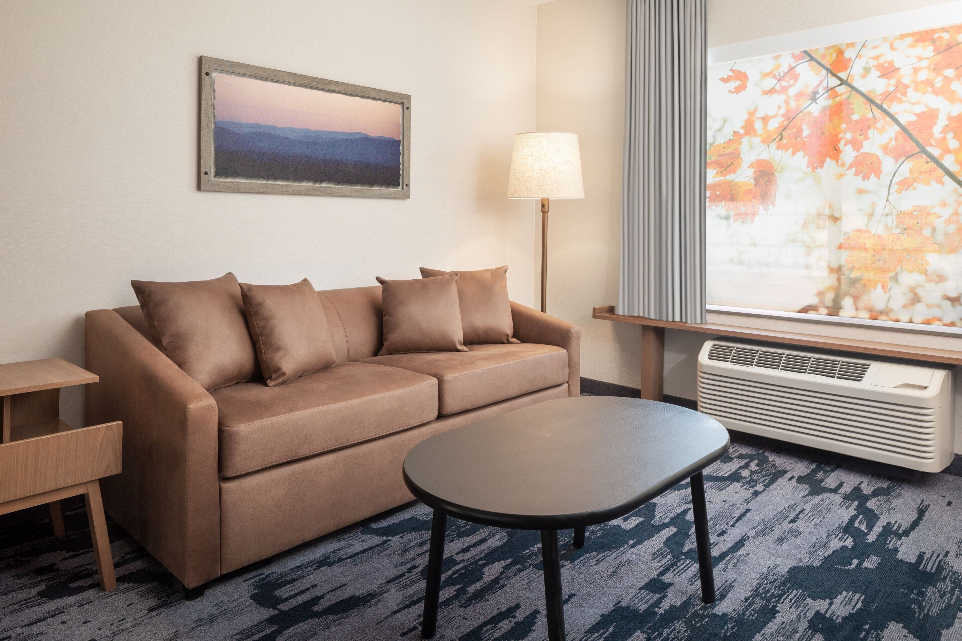 A hotel room with a brown sofa, a small round table, a floor lamp, a window with a view of autumn leaves, and a wall-mounted picture.
