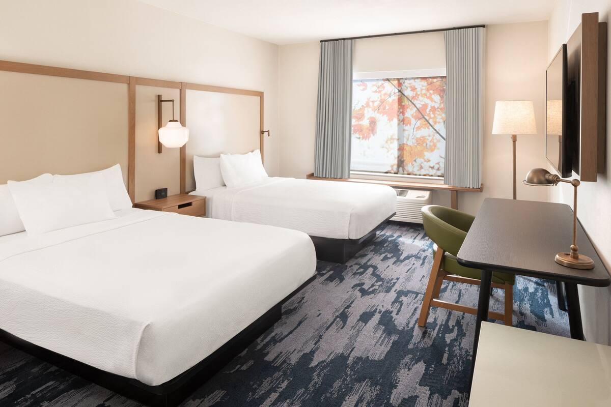 Une chambre d’hôtel avec deux lits, une petite table, une chaise, un bureau, une lampe et une fenêtre avec des rideaux.