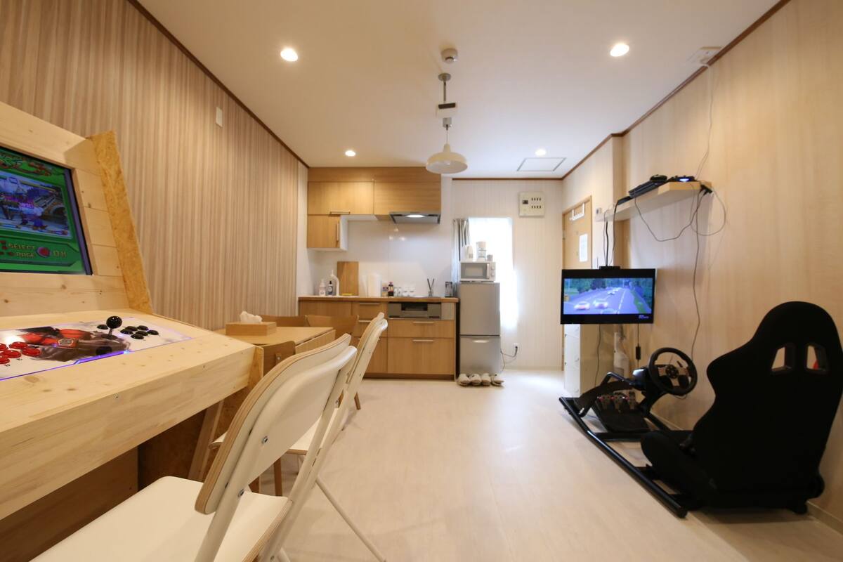 Een compacte leefruimte met een kitchenette, een gaming setup met monitor en stuur, en een aan de wand bevestigde televisie.