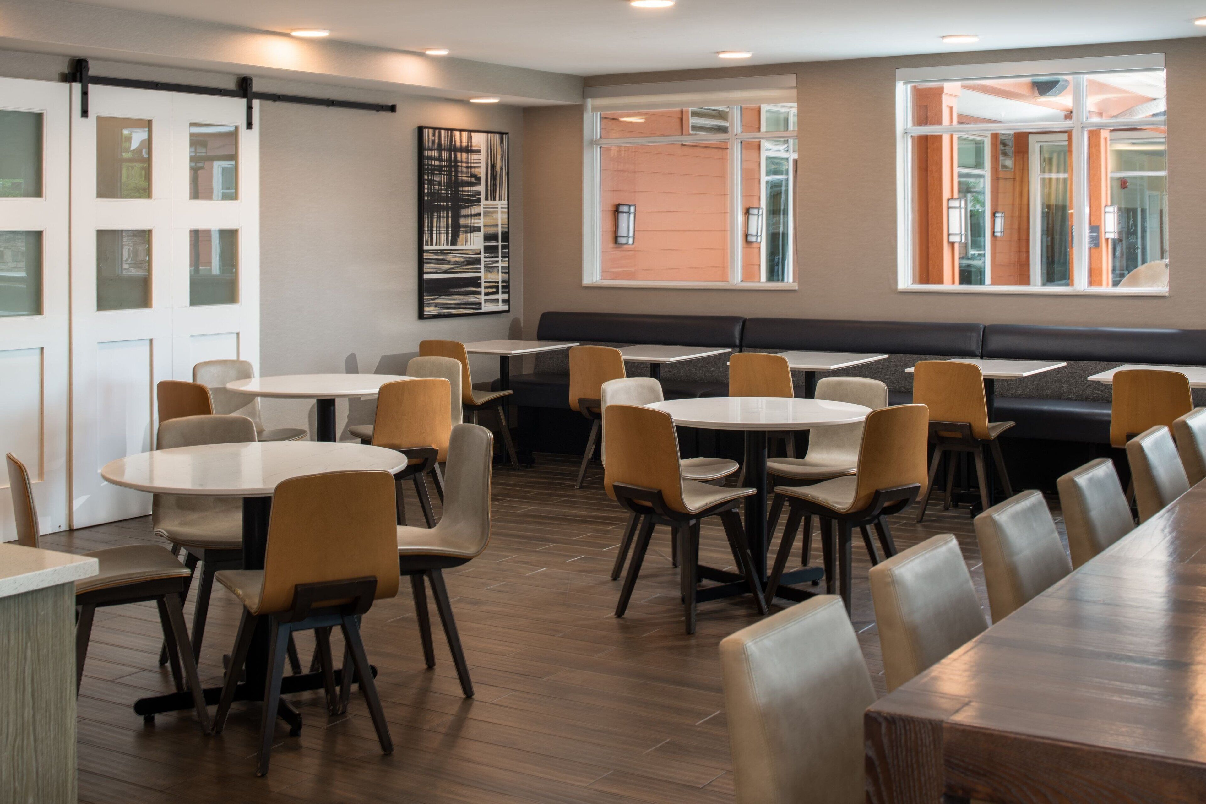 A modern dining area with round tables and booths, wooden flooring, and large windows offering views of the outside.
