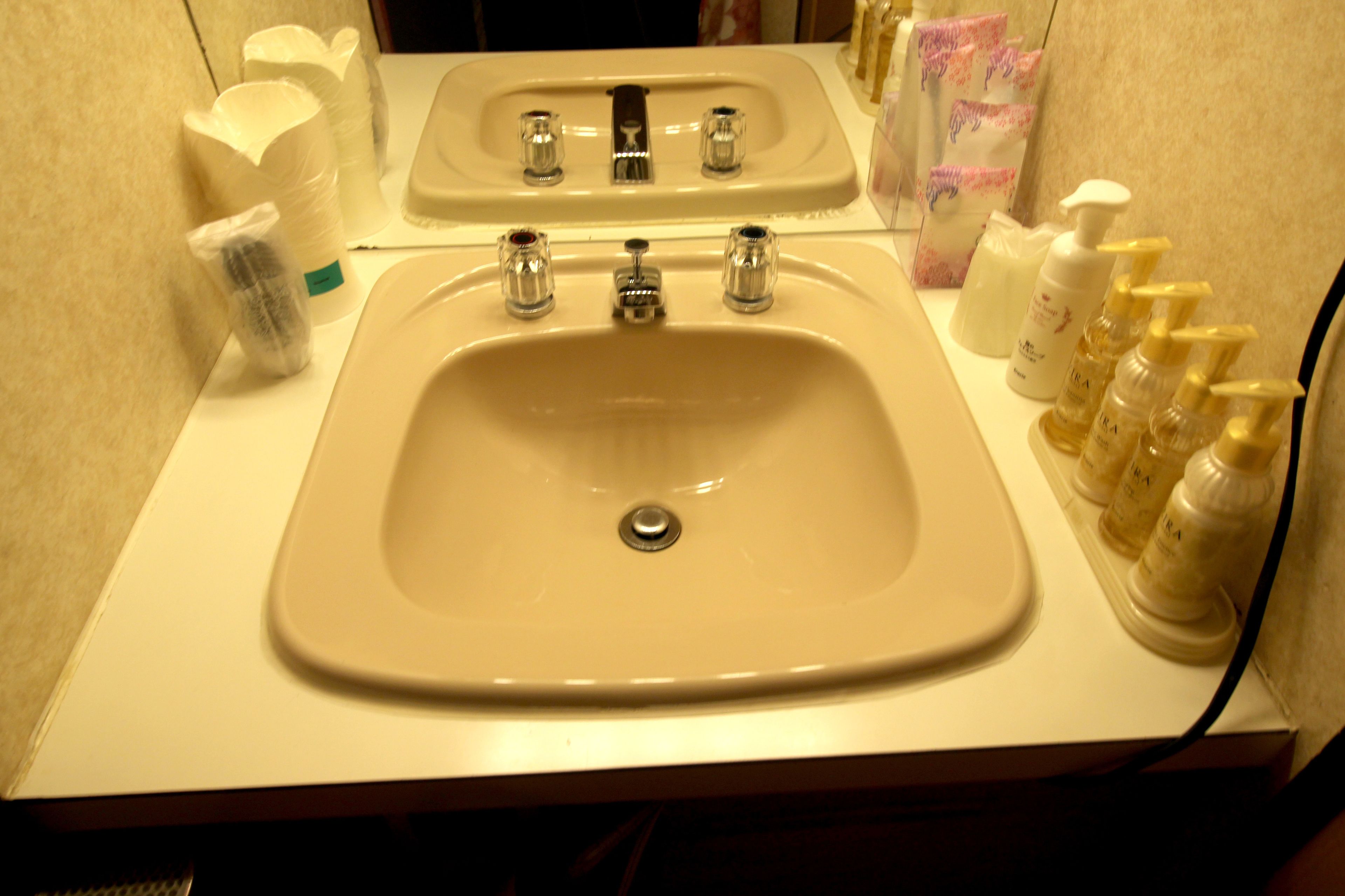 A bathroom with a sink and mirror, toiletries, and a towel dispenser.