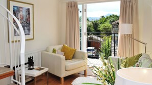 Ein modernes Wohnzimmer mit einem weißen Sofa, einem kleinen Tisch und Blick auf eine Stadtlandschaft durch große Fenster.