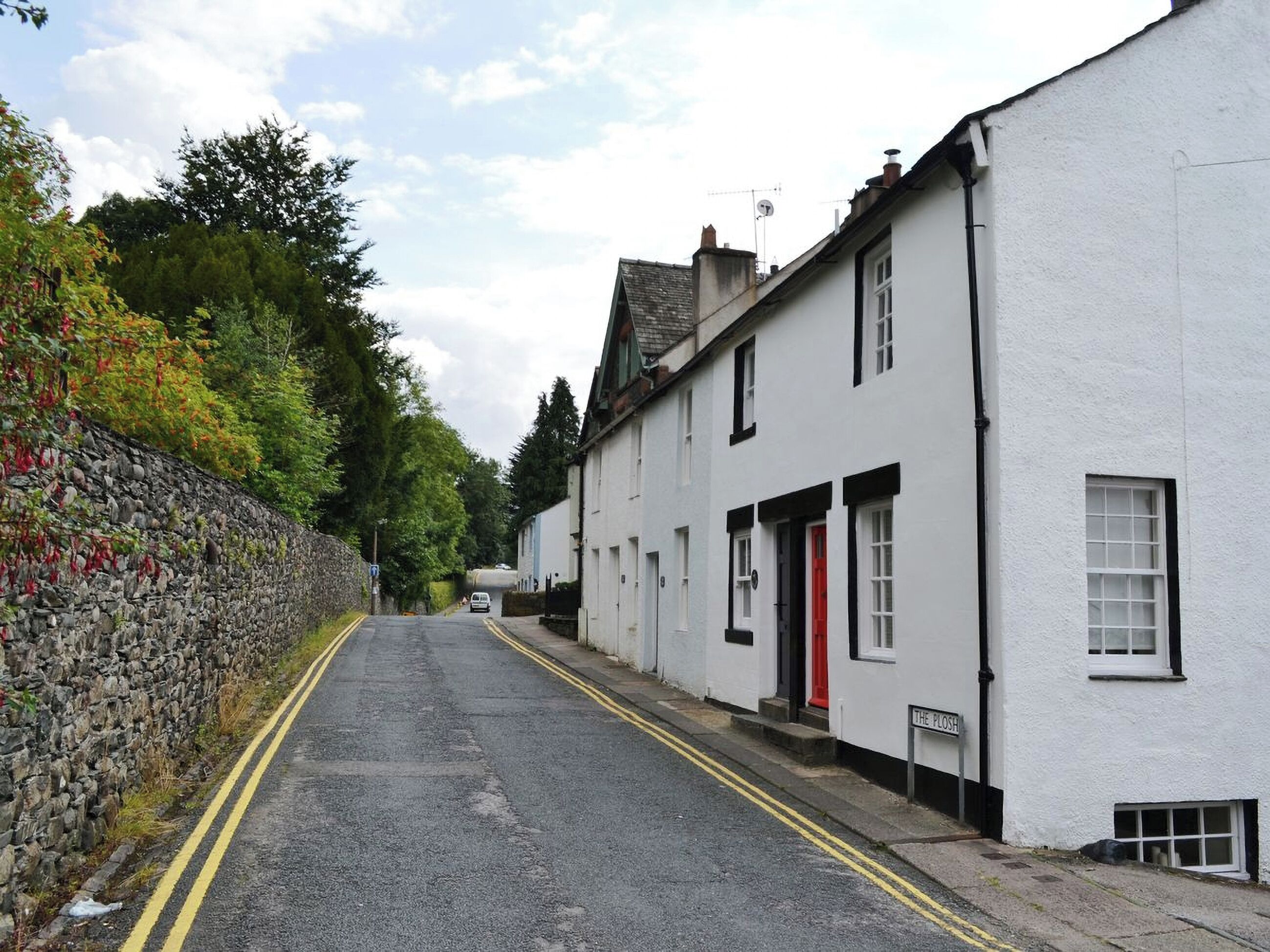 Uma rua estreita ladeada por edifícios brancos, um deles com uma porta vermelha com a placa 'The Plosh'.