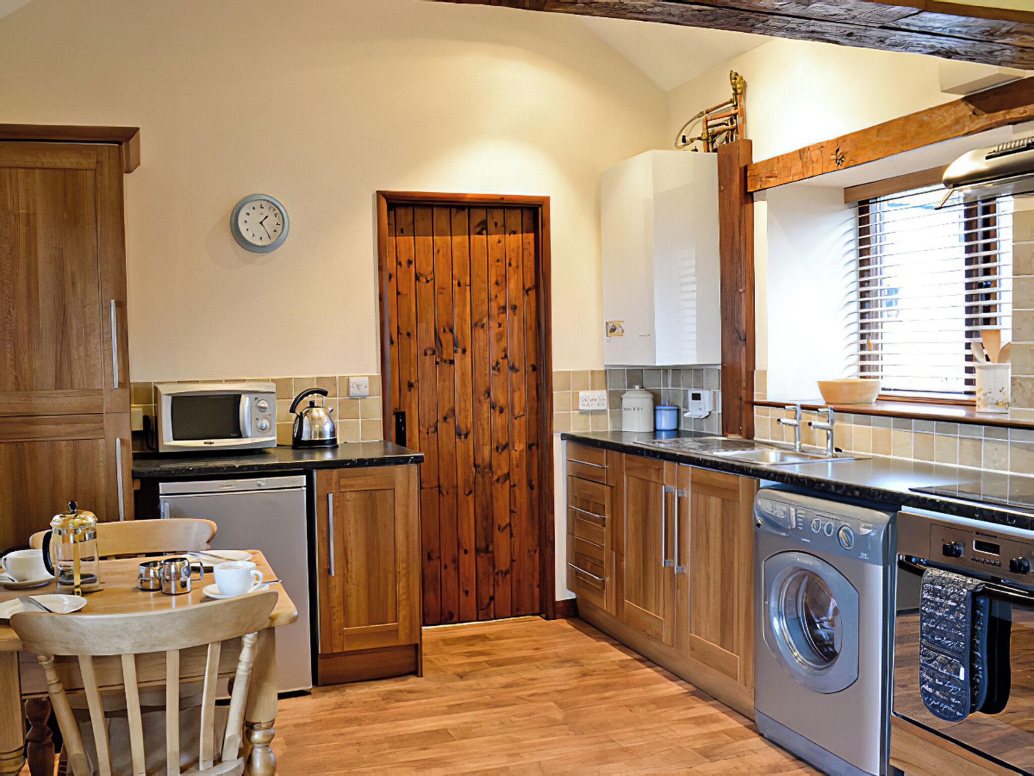 A compact kitchen with wooden cabinets, a microwave, a kettle, a washing machine, and a dining area with a table and chairs.
