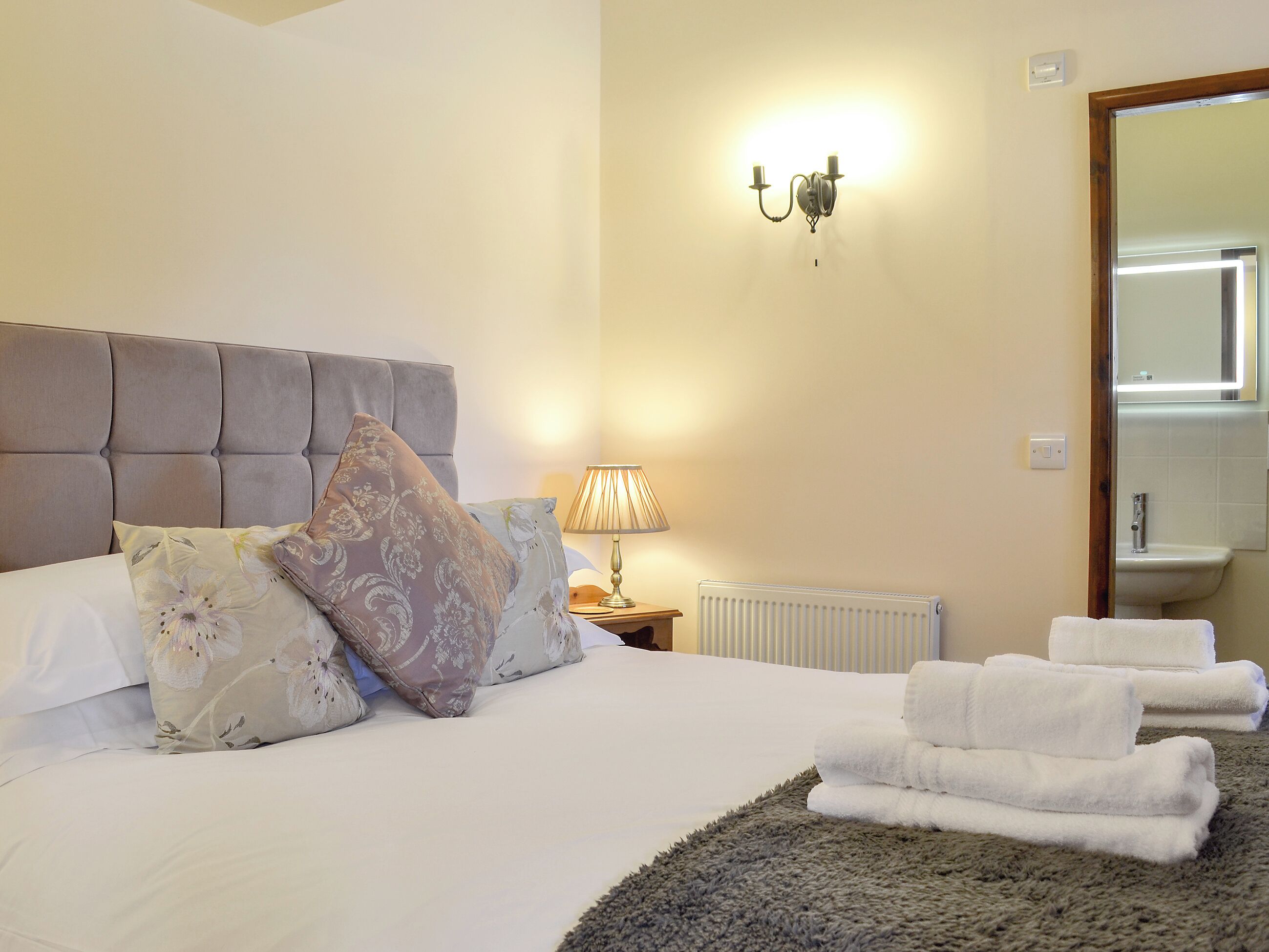 A neatly made bed with floral pillows, a bedside lamp, and a stack of towels.