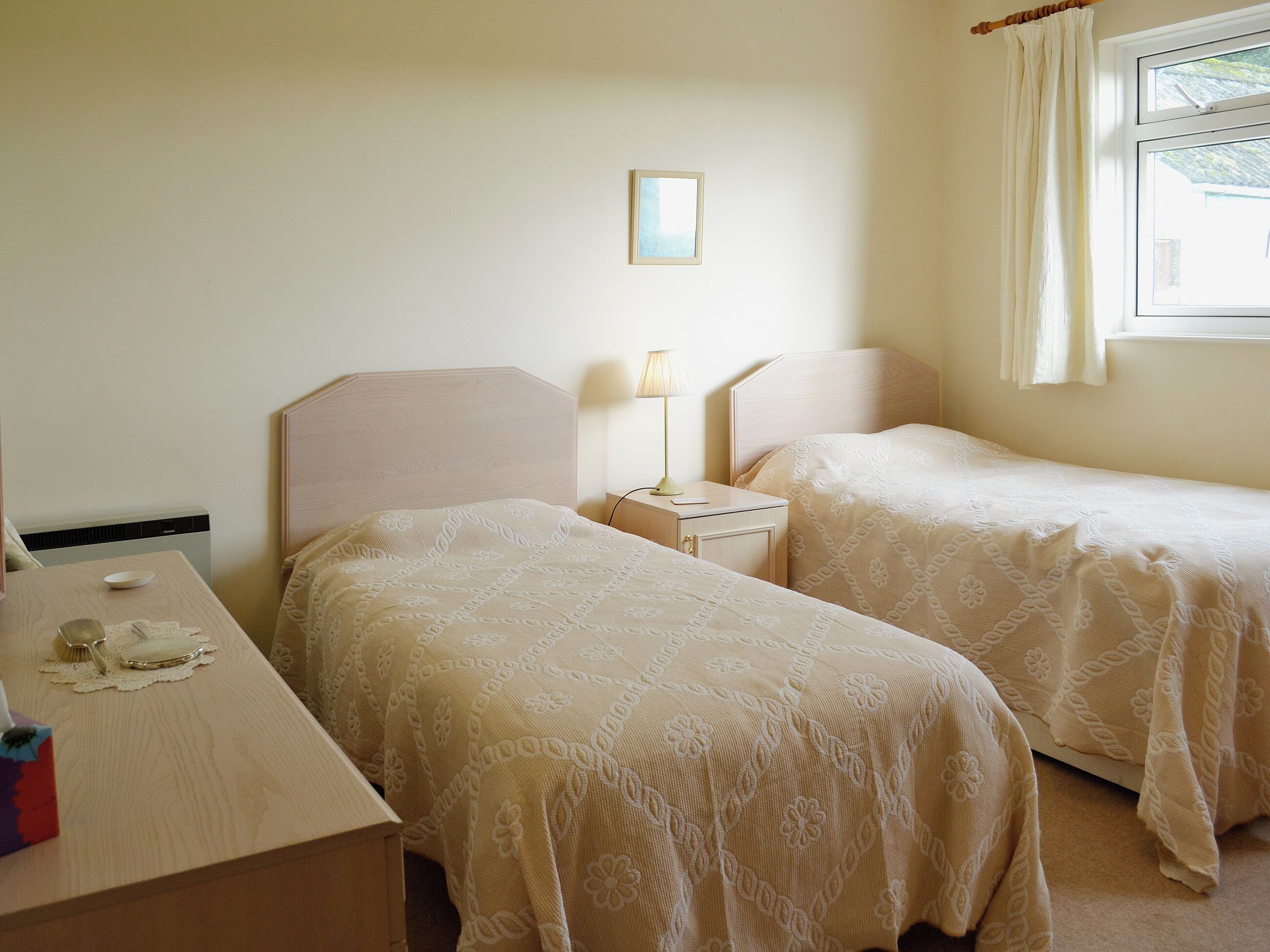 A room with two beds, a bedside table with a lamp, a small table with a tissue box, and a window with curtains.