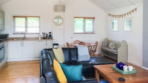 Amplia sala de estar con un sofá de cuero negro, una mesa de centro de madera y una mesa de comedor con sillas. Se ve una cocina con gabinetes blancos y un lavamanos al fondo.