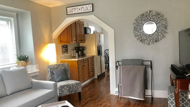 Sala de estar com sofá, poltrona estampada e mesa de centro. Há uma área de cozinha com armários de madeira e micro-ondas. Um espelho decorativo está pendurado na parede, e uma placa diz "HAPPY PLACE".