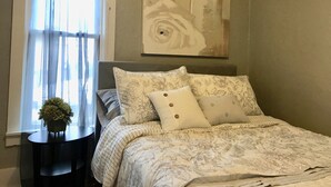 Quarto com cama, mesa de cabeceira, janela com cortinas leves e um quadro na parede.