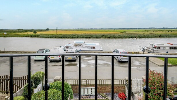 Een uitzicht op een gracht, rechts afgemeerd bootjes, een grasveld achterin en een gebouw met een hek in de voorgrond.
