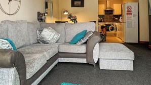 Ein modernes Wohnzimmer mit einem grauen Ecksofa, einem blaugrünen Teppich und einer im Hintergrund sichtbaren Küche.