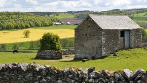Un edificio de piedra con techo inclinado, ubicado en un paisaje rural con campos verdes y un muro de piedra en primer plano.