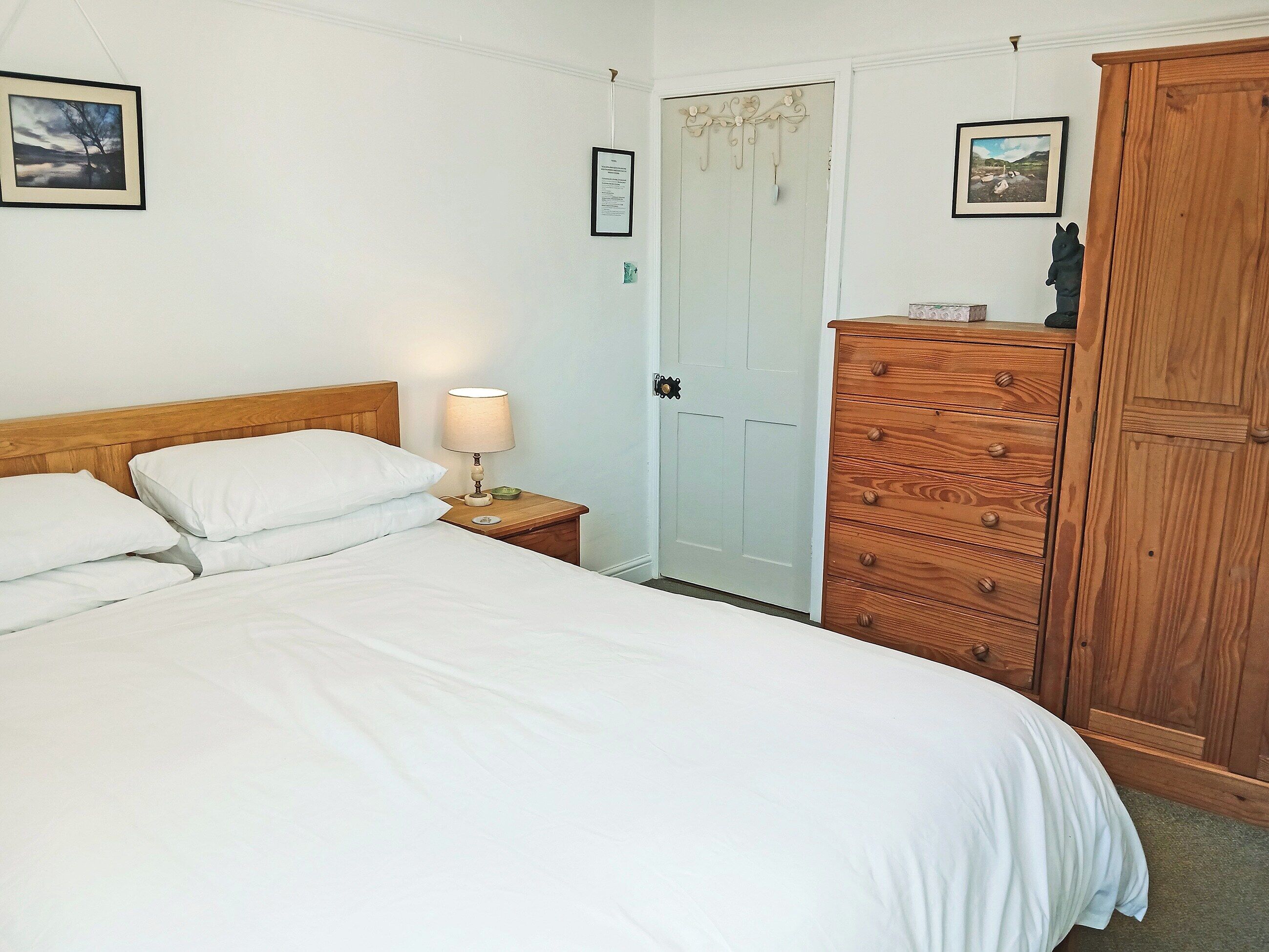 A neatly made bed with white linens, a wooden nightstand with a lamp, a wooden wardrobe, and two framed pictures on the wall.