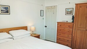 A neatly made bed with white linens, a wooden nightstand with a lamp, a wooden wardrobe, and two framed pictures on the wall.
