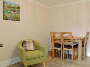 A room with a green armchair, a wooden dining table with chairs, and a landscape painting on the wall.