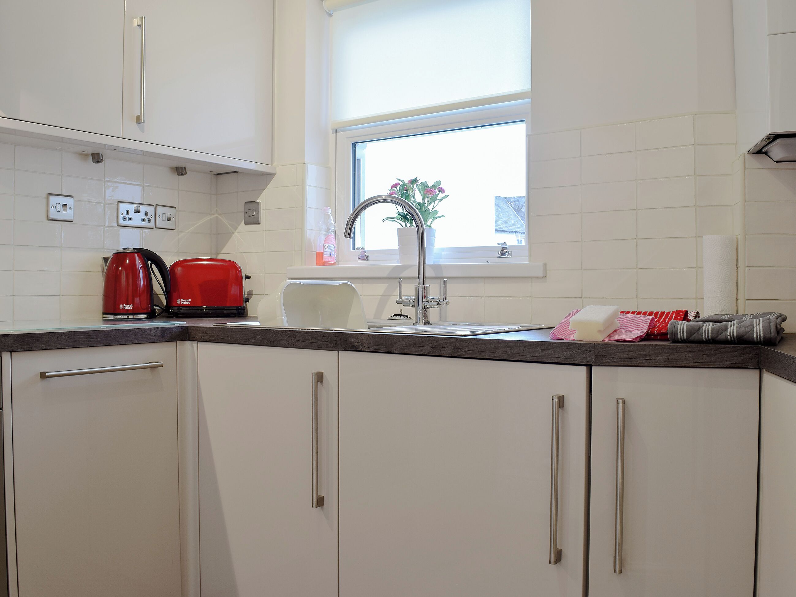 Eine Küche mit weißen Schränken, einem roten Wasserkocher, einem weißen Toaster, einer Spüle und einem Fenster mit Blick auf Grün.