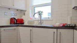 A kitchen with white cabinets, a red kettle, a white toaster, a sink, and a window with a view of greenery outside. - James' Retreat (was12 Elm Court) (Keswick)