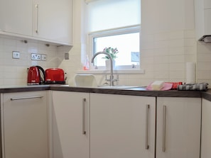 A kitchen with white cabinets, a red kettle, a white toaster, a sink, and a window with a view of greenery outside.