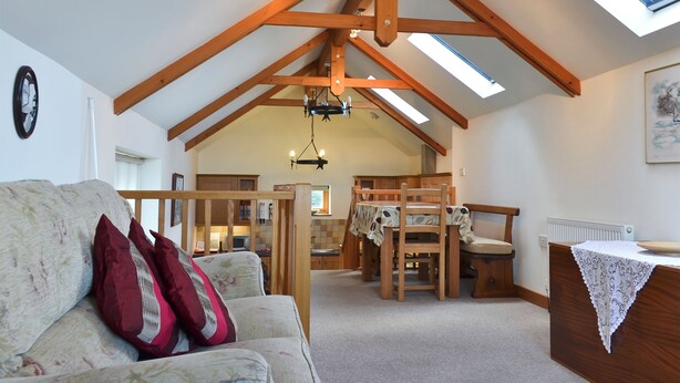 A spacious loft-style room with high ceilings, exposed wooden beams, a skylight, a cozy seating area with a sofa and cushions, a dining table with chairs, and a kitchen area in the background. - 1 bedroom accommodation in Port Isaac (Port Isaac)