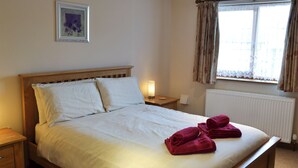 A neatly made bed with white linens and two red towels, a bedside lamp, a wooden nightstand, a framed picture on the wall, and a window with curtains. - 1 bedroom accommodation in Port Isaac (Port Isaac)