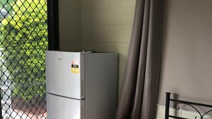 A compact room with a bed, a small wooden nightstand, a white refrigerator, and a window with a metal screen. - Close to amenities, NBN, Swimming pool, Netflix,  (Kirwan)