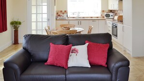 A modern living room with a black leather sofa, red and white cushions, a wooden dining table, and a kitchen in the background.