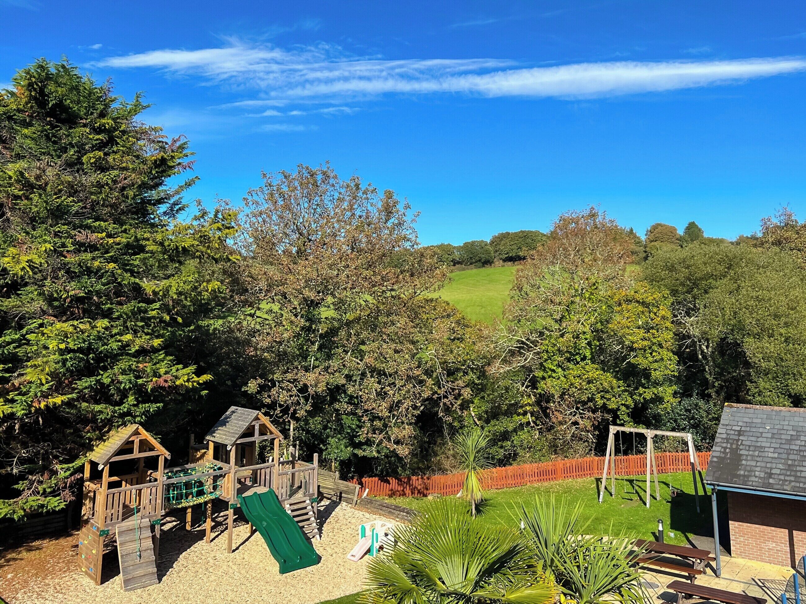 A playground with a slide and climbing wall, surrounded by trees and a grassy field.