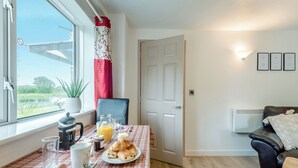 Un coin repas avec une table mise pour le petit-déjeuner, une vue sur un jardin, et un salon avec un canapé et un radiateur.
