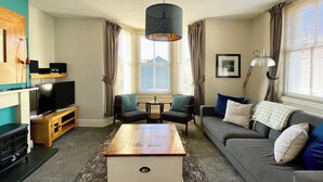 A living room with a wooden coffee table, grey sofa, and two armchairs. A television is on a wooden stand, and there's a window with curtains. - High Style Apartment (Keswick)