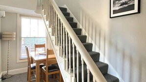 A staircase with white balusters and grey carpeted steps, a dining table with chairs, and a window with blinds. - High Style Apartment (Keswick)