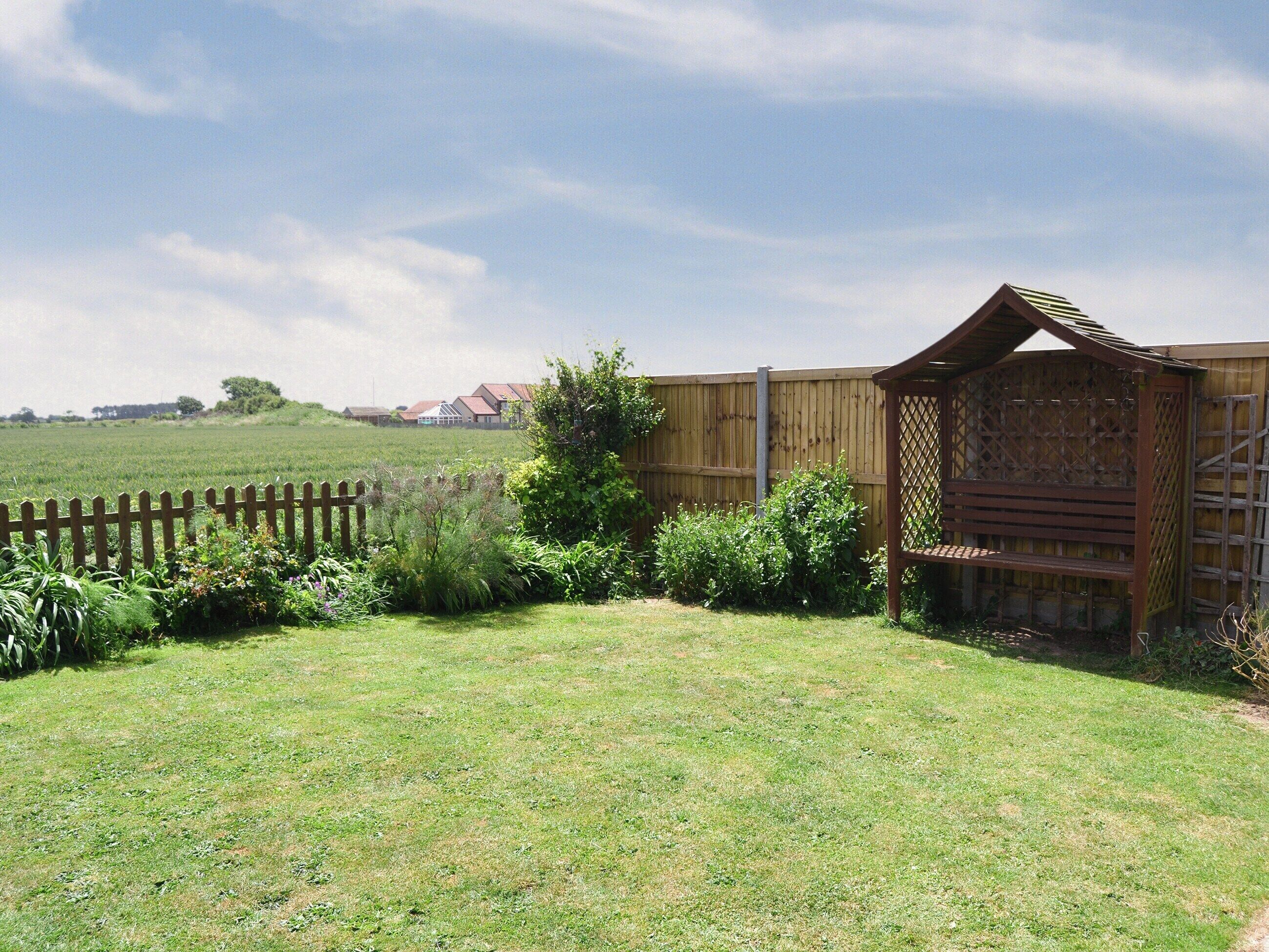 A grassy backyard with a wooden fence, a wooden structure with a lattice design, and a distant view of houses.