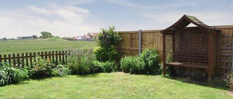 A grassy backyard with a wooden fence, a wooden structure with a lattice design, and a distant view of houses.