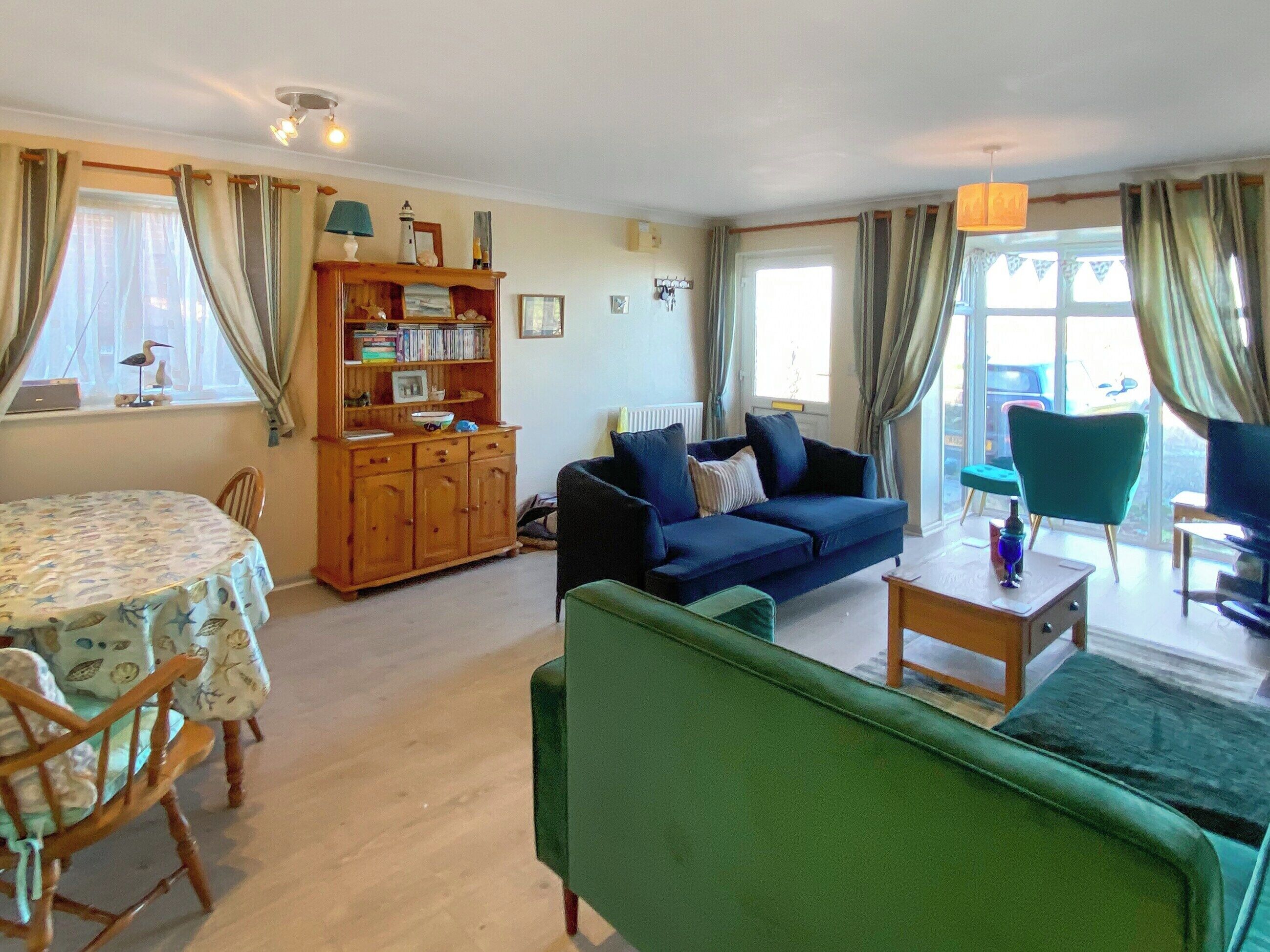 A cozy living room with a blue sofa, a green armchair, a wooden cabinet with books, and a dining table with chairs.
