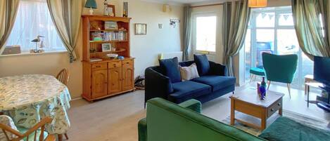 A cozy living room with a blue sofa, a green armchair, a wooden cabinet with books, and a dining table with chairs.