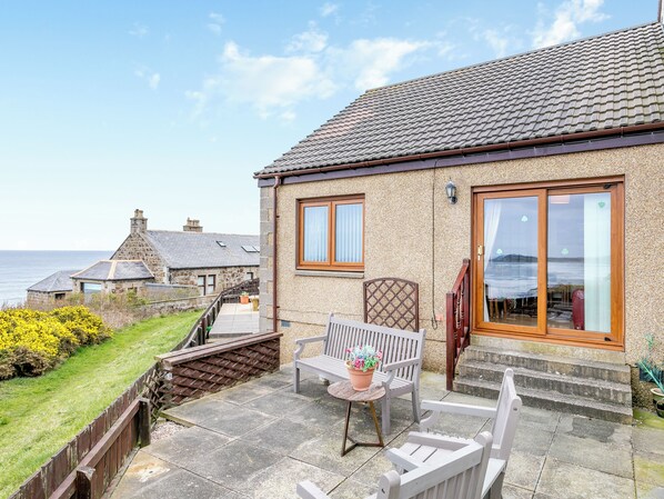 Une maison en pierre avec un toit en tuiles, de grandes fenêtres et une terrasse aménagée avec du mobilier d’extérieur.