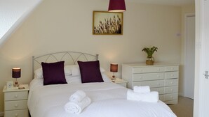 Una cama bien hecha con ropa blanca y dos almohadas moradas, una mesita de noche con una lámpara, un aparador con una planta encima y un cuadro en la pared.