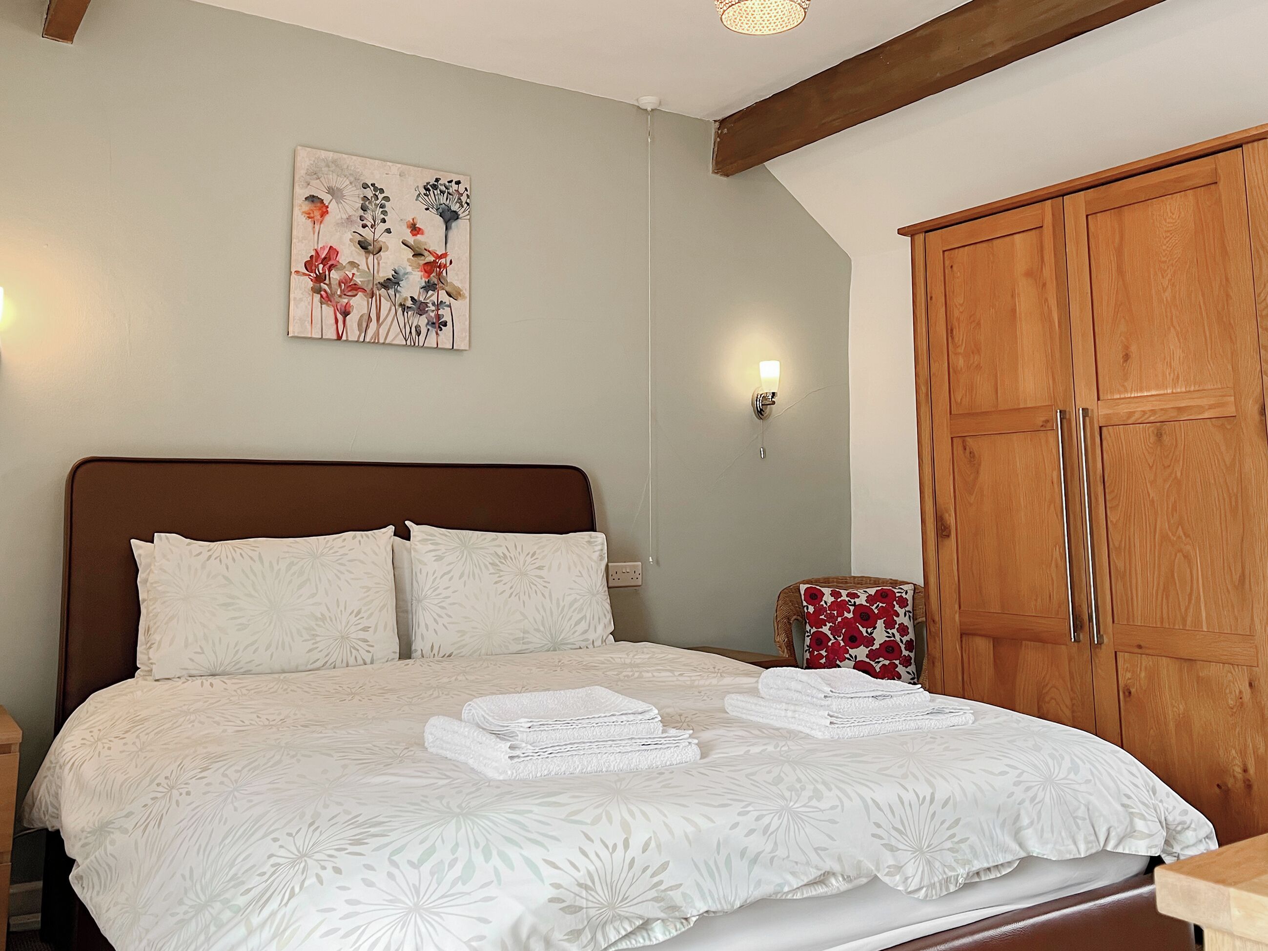 Una cama bien hecha con ropa blanca y motivos florales, un armario de madera y un cuadro de flores colgado en la pared.
