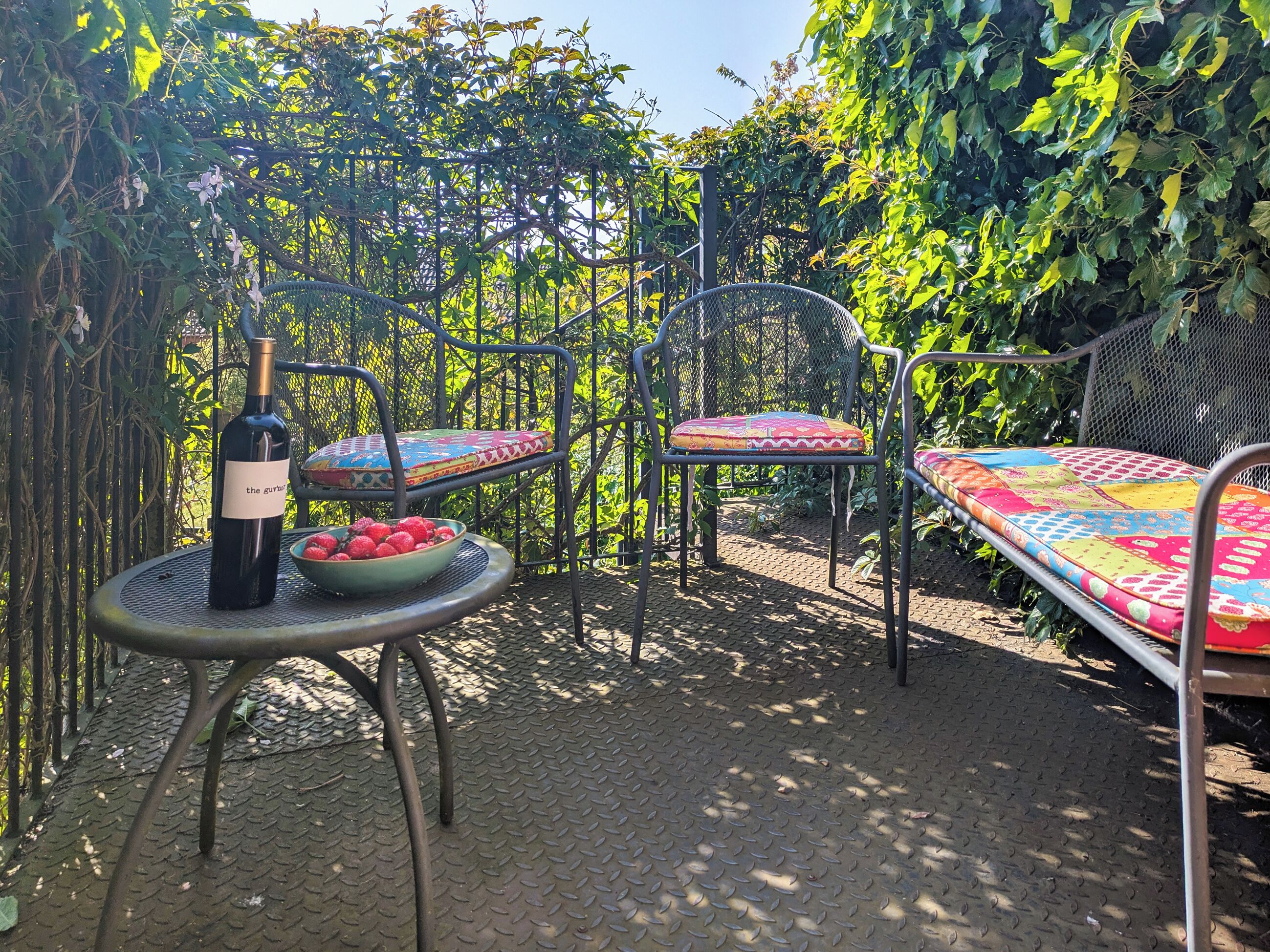 Een metalen tafel met een fles wijn en een schaal aardbeien, een kleurrijk kussen en een metalen stoel in een tuin.