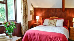 Een slaapkamer met een houten hoofdbord, een rode sprei, een nachtkastje met een lamp en een raam met gordijnen.