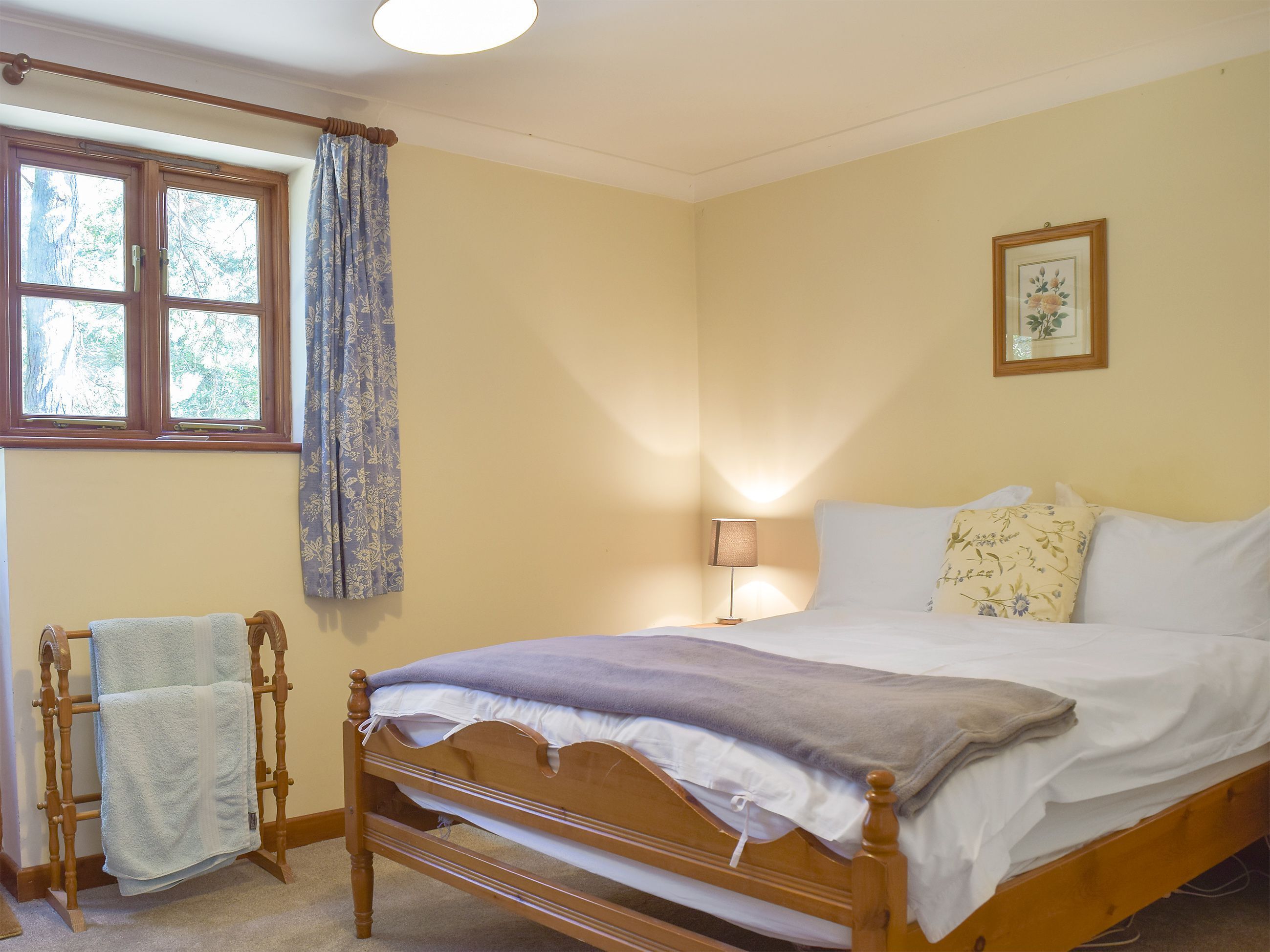 A bedroom with a wooden bed, a nightstand with a lamp, a window with curtains, and a towel rack.