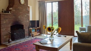 A living room with a brick fireplace, a television, a coffee table with a vase of sunflowers, and a large window with curtains. - 3 bedroom accommodation in Bromeswell, Woodbridge (Bromeswell, Woodbridge)