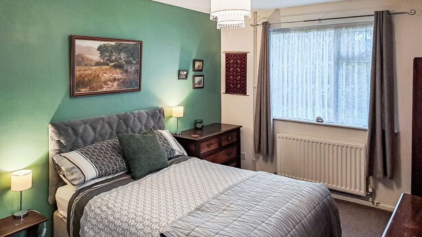 A bedroom with a bed, bedside lamps, a nightstand, a dresser, a window with curtains, and a hanging light fixture.